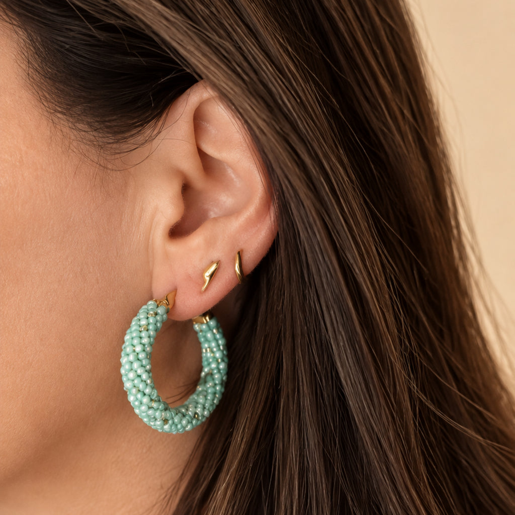 Créole en acier inoxydable portée à l’oreille, tissée à la main en perles Miyuki vertes, blanches et dorées, diamètre environ 3,5 cm.
